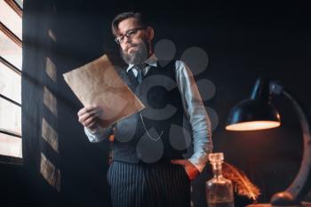 Seriuos bearded writer in glasses reads handwritten text against the window with sunlight. Retro typewriter, feather, crystal decanter, books and vintage lamp on the desk