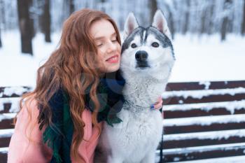 Happy woman sitting on the bench with siberian husky, snowy forest on background. Cute girl hugs with charming dog. Real friendship with pet