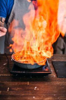 Male person against frying pan with fire, cooking sea bass fish fillet. Seafood preparation