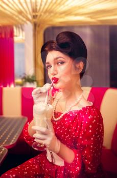 Sexy pin up girl with make-up drinks milkshake through a straw in retro cafe, popular american fashion 50s and 60s. Red dress with polka dots, vintage style