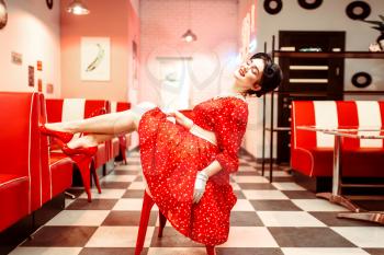 Sexy pin up girl with makeup posing on the chair in retro cafe, 50 american fashion. Red shoes and dress with polka dots, vintage style