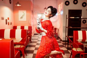 Pinup girl with make-up drinking popular carbonated drink in retro cafe, 50 american fashion. Red dress with polka dots, vintage style