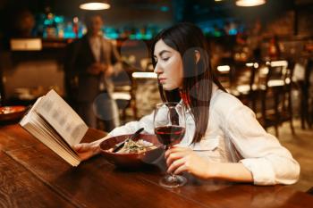 Young woman drinks red wine and reading a book at wooden bar counter. Female customer relaxing in pub