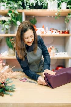 Smiling seller packing gift box in wrapping paper, decoration process. Woman wraps present on the table, decor procedure