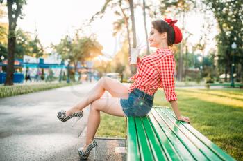 Sexy pinup girl sitting on brench and holds cardboard cup with a straw, city park on background. Vintage american fashion. Attractive woman in pin up style
