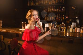 Portrait of young woman in red dress sitting at the bar counter. Beautiful lady with cocktail in hand in club