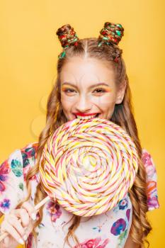 Beautiful young woman with playful look eating huge candy and smiling. Stylish girl with blonde curly hair. Portrait of attractive lady with big lollypop, yellow wall on background.