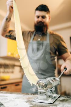 Bearded chef in apron works with dough in pasta machine on wooden kitchen table. Homemade spaghetti cooking process