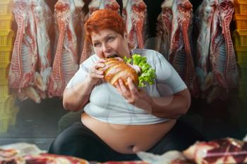 Fat woman eating big burger against meat carcasses. Overweight concept. Female obesity, bulimic. Unhealthy food eating