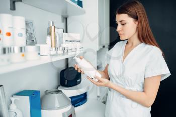 Young female beautician with bottle of cream in hands, cosmetology cabinet. Doctor in white uniform in spa salon, beauty medicine