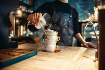 Male barista pours black coffee on a stack of cups, cafe counter and espresso machine on background. Barman works in cafeteria, bartender occupation