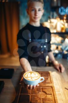 Male barista shows cup of coffee with foam drawing, top view, wooden cafe counter on background. Professional cappuccino preparation by barman in cafeteria, bartender art occupation
