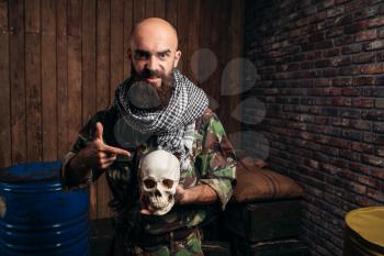 Terrorist in uniform holds human skull in hands, male mojahed. Terrorism and terror horror, bearded soldier in khaki camouflage, barrels of fuel or chemicals on background