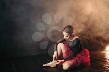 Ballet dancer in red dress sitting on the stage floor in theatre and tying pointe shoes. Elegance ballerina training in class