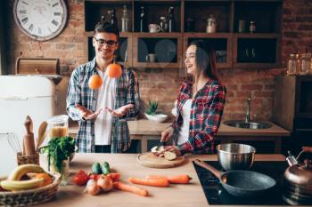 Young husband juggles oranges on the kitchen while wife cooking fruit breakfast. Man and woman preparing vegetable salad, happy family together