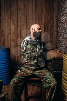 Captive soldier sitting tied to a chair, horror of war. Terrorism and terror, military man in khaki camouflage, barrels of fuel or chemicals on background