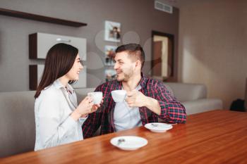Young happy love couple drinks coffee at wooden table in the morning. Modern apartment interior on background.