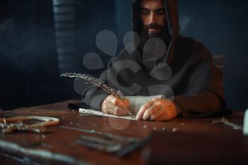 Medieval monk sitting at the table and writes with a goose feather, top view, secret scripture. Mysterious friar in dark cape. Mystery and spirituality