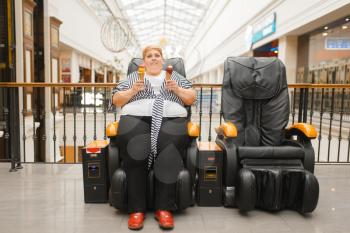 Fat woman with ice cream sitting in a massage chair in mall. Overweight female person poses in a leather armchair in shopping center, obesity problem