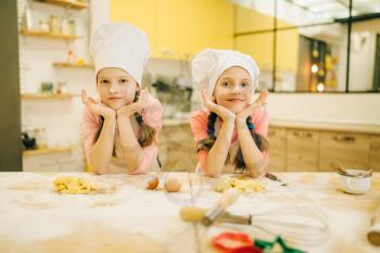 Two little girls cooks in caps are sitting at the table, cookies preparation on the kitchen. Kids cooking pastry, children chefs preparing cake