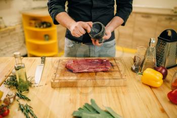 Male person sprinkles raw meat with seasonings, kitchen interior on background. Chef cooking tenderloin with vegetables, spices and herbs