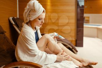Sexy girl in bathrobe and towel on the head applying cream in spa chair, body care. Relaxation leisure, healthy lifestyle, attractive woman resting in armchair, beauty salon