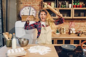 Furious housewife in an apron with frying pan and rolling pin in hands, kitchen interior on background. Female cook prepares homemade cake. Domestic pie preparation