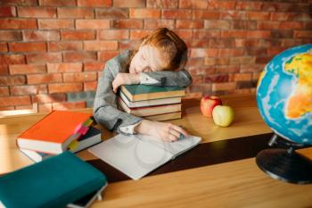 Tired female pupil asleep on the stack of textbooks at the table with opened notebook. Schoolgirl sleeping at the desk in the school