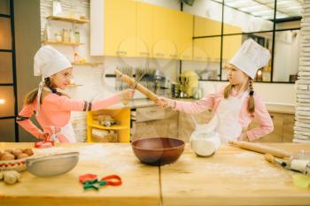 Two girls cooks in caps fights on the kitchen. Kids cooking pastry, little chefs holds rolling pin and whisk for whipping