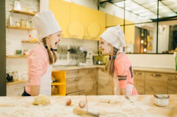 Two little girls chefs are plays and having fun, cookies preparation on the kitchen. Kids cooking pastry, children cooks preparing cake