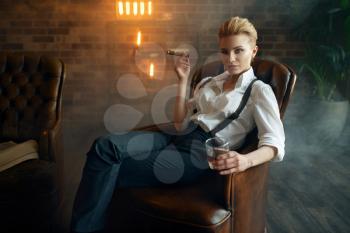 Woman in shirt and trousers sitting in leather chair with whiskey and cigar, retro fashion, gangster style. Vintage business lady in office with brick walls