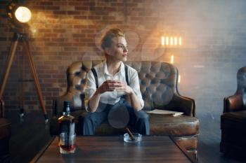 Woman in shirt and trousers sitting at the table with whiskey and cigar, retro fashion, gangster style. Vintage business lady