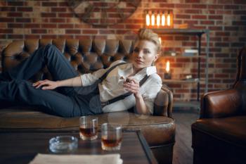 Woman in strict clothes lying on leather couch and smoking cigar, retro fashion, gangster style. Vintage business lady in office with brick walls
