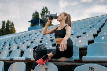 Female runner in sportswear sitting on tribune and drinks water, training on stadium. Woman doing stretching exercise before running on outdoor arena