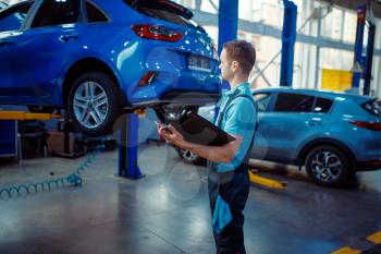 Repairman with a checklist stands at vehicle on lift, car service station. Automobile checking and inspection, professional diagnostics and repair