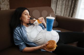 Pregnant woman with belly drinks and eats junk food at home. Pregnancy, gluttony in prenatal period. Expectant mom resting in living room