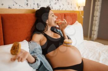 Pregnant woman with belly eats sweet cakes in bed at home. Pregnancy, gluttony in prenatal period. Expectant mom resting in bedroom