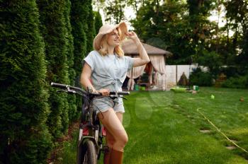Attractive female gardener poses on bicycle in the garden. Woman on cycle outdoor, gardening hobby, florist lifestyle and leisure
