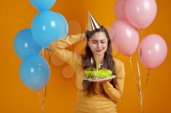 Pretty woman in cap holding birthday cake with firework, yellow background. Smiling male person got a surprise, event celebration, balloons decoration