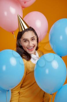 Funny woman in cap, yellow background. Pretty female person got a surprise, event or birthday celebration, balloons decoration