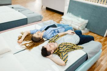 Young love couple lying in bed in furniture store showroom. Man and woman looking samples for bedroom in shop, husband and wife buys goods for modern home interior