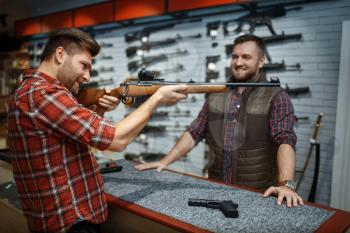 Man aims with new rifle, seller at counter in gun shop. Male person buying weapon in store, hunting and sport shooting hobby