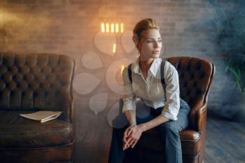 Elegance business woman in strict clothes poses on leather couch in studio, retro fashion, gangster style, mafia.