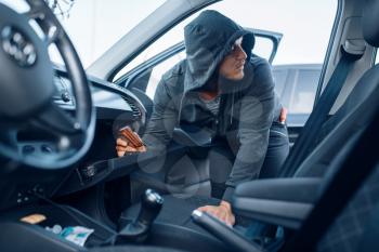Car robber takes the wallet from the glove compartment, criminal lifestyle, stealing. Hooded male bandit opening vehicle on parking. Auto robbery