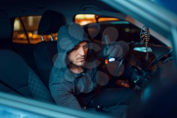Professional car thief with flashlight, criminal lifestyle, stealing. Hooded male robber looking for goods in vehicle on parking, steal. Auto robbery, automobile crime
