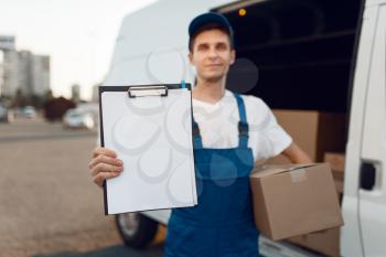 Deliveryman in uniform holding parcel and notebook, carton boxes in the car, delivery service. Man standing at cardboard packages in vehicle, male deliver, courier or shipping job