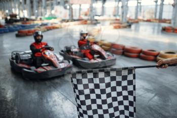 Two kart racers on start line, checkered flag, front view, karting auto sport indoor. Speed race on close go-cart track with tire barrier. Fast vehicle competition, high adrenaline leisure
