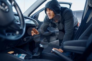 Car robber takes the wallet from the glove compartment, criminal lifestyle, stealing. Hooded male bandit opening vehicle on parking. Auto robbery, automobile crime