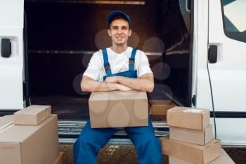 Deliveryman in uniform sitting between parcels and carton boxes, delivery service, delivering occupation. Man poses at cardboard packages in vehicle, male deliver, courier or shipping job