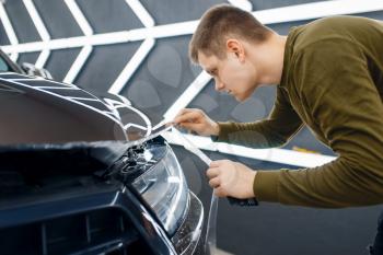 Male worker cuts transparent protection film on car hood. Installation of coating that protects the paint of automobile from scratches. New vehicle in garage, autotuning